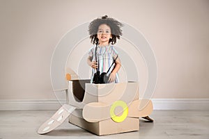 Cute African American child playing with cardboard plane and binoculars near wall