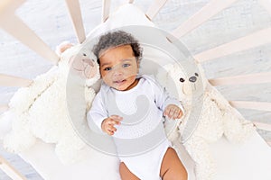 Cute African-American little baby in white sleeping bed with bear toys