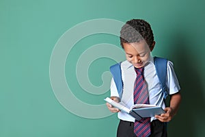 Cute African-American schoolboy reading book on color background