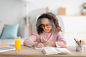 Cute african american schoolgirl in glasses writing in notepad, doing homework or writing essay, studying at home