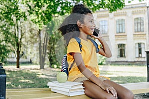 African american schoolgirl talking on smartphone while sitting on bench near books and apple