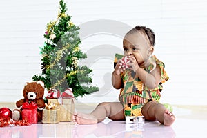 Cute African baby kid in colorful dress sits near decorative Christmas tree and gift box present on white wall, little girl child