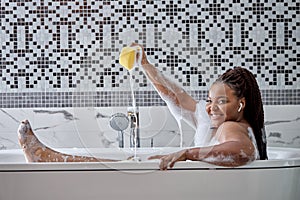 cute african black lady in bathtub with foam, playing splashing with water holding sponge