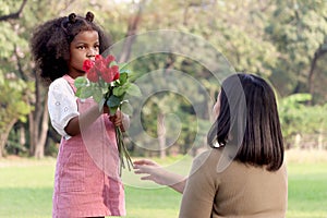 Cute African girl with black curly hair giving red rose flower to her mother while having a picnic at green garden park,