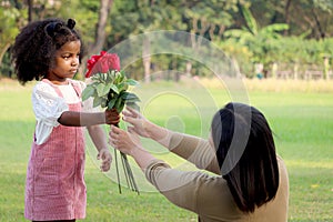 Cute African girl with black curly hair giving red rose flower to her mother while having a picnic at green garden park,