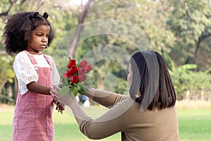 Cute African girl with black curly hair giving red rose flower to her mother while having a picnic at green garden park,