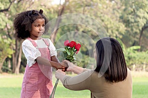 Cute African girl with black curly hair giving red rose flower to her mother while having a picnic at green garden park,