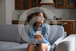 Cute African girl sit on sofa with smartphone staring aside