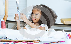 Cute African little girl holding scissor cutting clothing, design on paper at tailor room, cozy home as her career dream, smiling