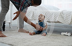 Cute African toddler baby smiling to father who standing and bow down give her sweets while sitting on carpet on floor at bedroom