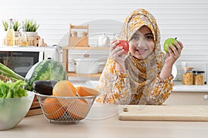 Cute Arab muslim kid girl wearing hijab smiling and holding red and green apples in kitchen at home with many fruits