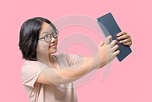 Cute asia girl wearing glasses takes a selfie of herself with a tablet on pink background