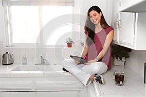 Cute asian american female sitting cozy with tablet and tea on kitchen counter, morning ritual, copy space