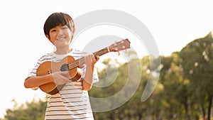 Cute Asian boy holding Ukulele with a big smile feeling happy and confident to play music in the park, carefree mind, concept