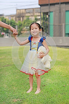 Cute Asian child girl dressed a fantasy outfit holding teddy bear and unshod walking on green grass garden with mother hand in