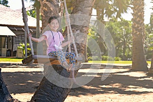 Cute Asian child girl sitting on wooden swing at resort in Thailand, swinging under palm tree, sand beach, hang medical mask on