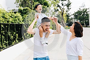 Cute Asian father piggybacking his son along with his wife in the park. Excited family raising hands together with happiness.