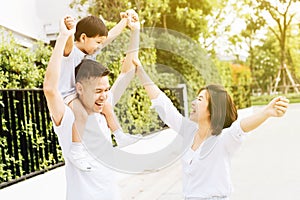 Cute Asian father piggybacking his son along with his wife in the park. Excited family raising hands together with happiness.