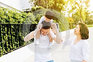 Cute Asian father piggybacking his son along with his wife in the park. Excited family spending time together with happiness.