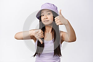 Cute asian girl asking your opinion, showing thumbs up and down and looking at camera, need advice, standing over white