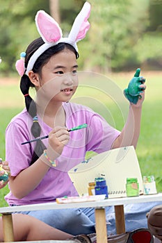 Cute Asian girl with bunny ears painting eggs with paintbrush while sitting on green grass meadow in nature garden. Kid