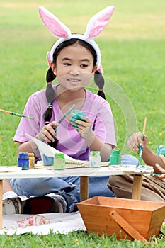 Cute Asian girl with bunny ears painting eggs with paintbrush while sitting on green grass meadow in nature garden. Kid