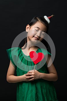 Cute asian girl child dressed in a green dress holding a Christmas ornament and a heart stick on a black background