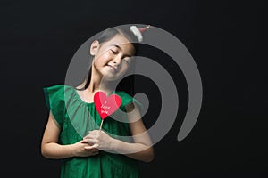 Cute asian girl child dressed in a green dress holding a Christmas ornament and a heart stick on a black background