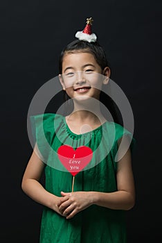 Cute asian girl child dressed in a green dress holding a Christmas ornament and a heart stick on a black background