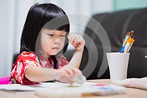 A cute Asian girl is holding a cotton ball dipped in paint and touching the paint onto a piece of art paper.