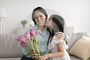 Cute Asian girl kissing cheerful grandmother, giving her flowers for Women& x27;s Day, greeting with birthday at home