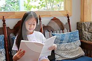 Cute Asian girl reading a book while sitting on wood chair in living room