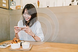 Cute Asian girl taking photo of dessert at coffee shop. Leisure activity or mobile phone photography, food photo concept.