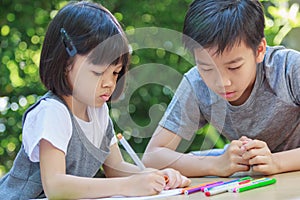 Cute Asian girls and boys, aged 4 to 10, sit outdoors in the garden, the brother is teaching her sister to write and draw with joy