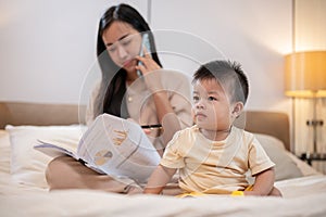 A cute Asian little boy is playing toys in bed while his mother is working and talking on the phone