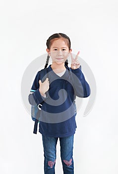 Cute asian little child girl in casual school uniform with backpack showing finger fight or victory sign on white background.