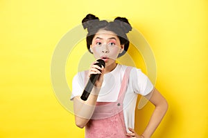 Cute asian teen girl with bright makeup, singing in microphone karaoke, standing against yellow background