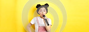 Cute asian teen girl with bright makeup, singing in microphone karaoke, standing against yellow background