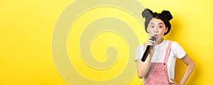 Cute asian teen girl with bright makeup, singing in microphone karaoke, standing against yellow background