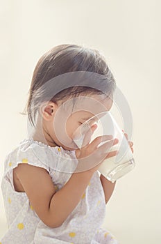 Cute asian toddler girl drinking milk from big glass on white ba
