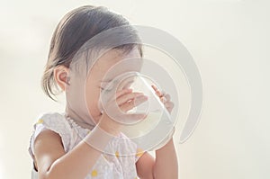 Cute asian toddler girl drinking milk from big glass on white ba