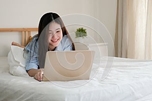 Cute asian woman in blue shirt using computer laptop for work at home  on bed.Brightly smile woman working in the bedroom.