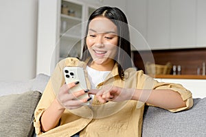Cute asian woman looking at the smartphone in her hands. Female using phone while sitting down on the sofa