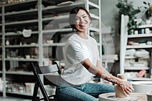A cute Asian woman sits at a potter& x27;s wheel in a hobby studio and makes a clay vase. Beautiful smile, white teeth