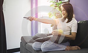 Cute Asian woman sitting on sofa with relax and cozy gesture holding remote control aiming to tv, watching television and smile