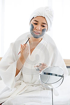 A cute Asian woman wearing spa dress and covered head with white towel using brush to add treatment mud or cream on her own face