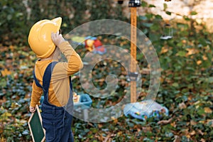 Cute asian young kid builder boy wearing protective yellow helmet on background construction cranes on site. Future