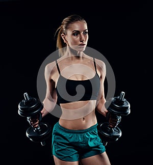 Cute athletic model girl in sportswear with dumbbells in studio against black background. Ideal female sports figure.