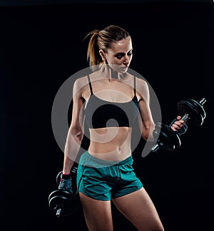 Cute athletic model girl in sportswear with dumbbells in studio against black background. Ideal female sports figure.