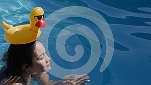 A cute attractive female person swimming in a swimming pool with a funny inflatable yellow duck on head. Asian girl